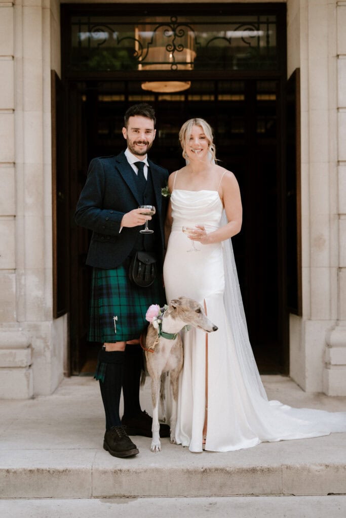 A couple stands in front of a building, dressed in wedding attire. The man wears a dark jacket and a green tartan kilt, while the woman wears a white gown. They smile and hold drinks, accompanied by a greyhound with a pink rose on its collar, captured beautifully by their Bethnal Green wedding photographer. Image by Pearce Wedding Photography.