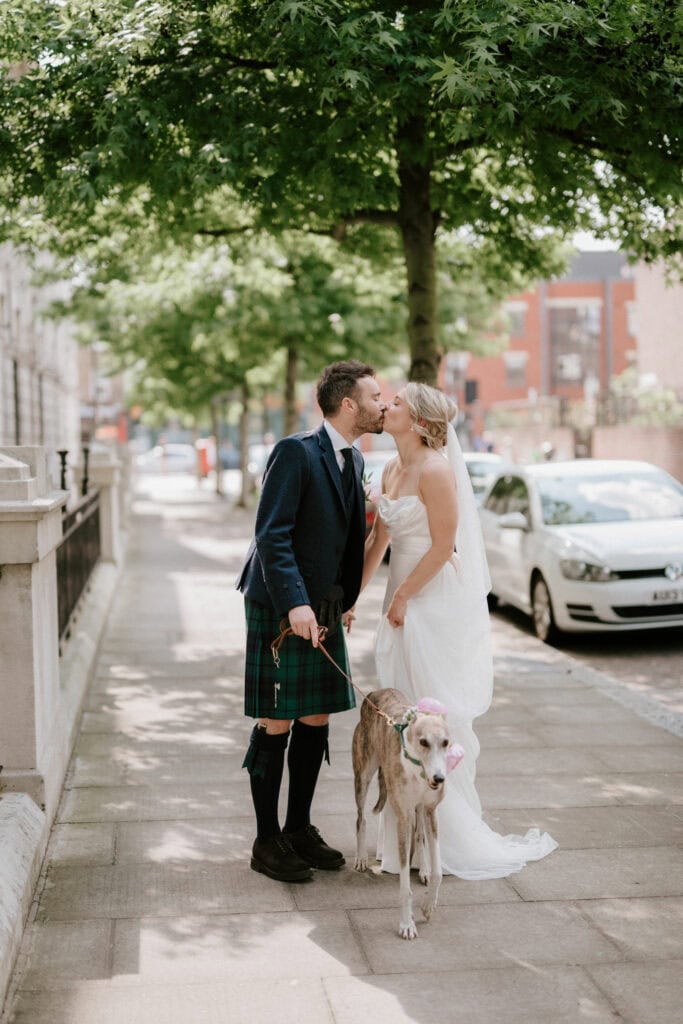 A bride and groom kiss on a city sidewalk under a tree. The groom wears a kilt, navy jacket, and black knee socks. The bride is in a white strapless wedding gown with a veil. They are holding a leash attached to a greyhound wearing a floral headpiece, beautifully captured by their Bethnal Green wedding photographer. Image by Pearce Wedding Photography.