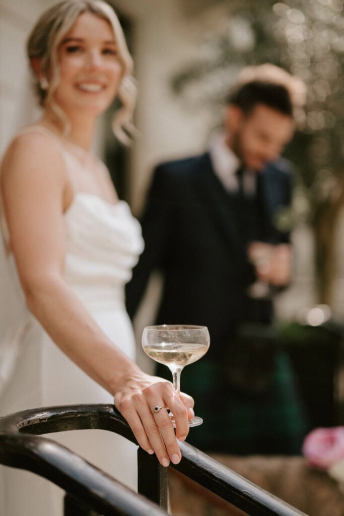 A woman in a white dress, holding a glass of champagne, stands in the foreground, smiling. A man in a suit stands blurred in the background, also holding a drink. The setting appears to be outdoors with greenery around—a perfect moment captured by a Bethnal Green wedding photographer. Image by Pearce Wedding Photography.