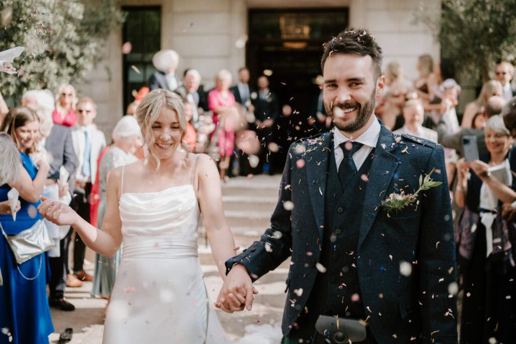 A bride in a white dress and a groom in a dark suit walk hand-in-hand down an outdoor aisle, surrounded by cheering guests. Confetti fills the air, and both are smiling joyfully. The scene is lively and celebratory as the Bethnal Green wedding photographer captures every moment on camera. Image by Pearce Wedding Photography.