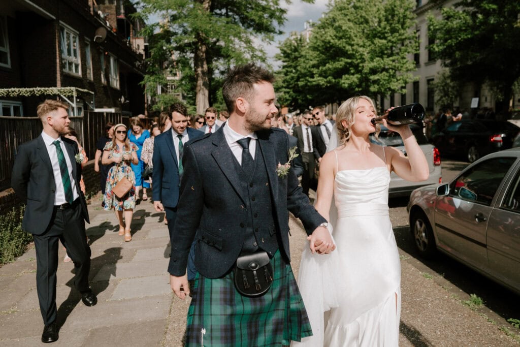 A bride in a white dress drinks from a bottle while holding hands with the groom, who is wearing a kilt and suit jacket. Captured beautifully by their Bethnal Green wedding photographer, they walk on a sunny street followed by cheerful friends and family in formal attire, with trees and parked cars lining the way. Image by Pearce Wedding Photography.