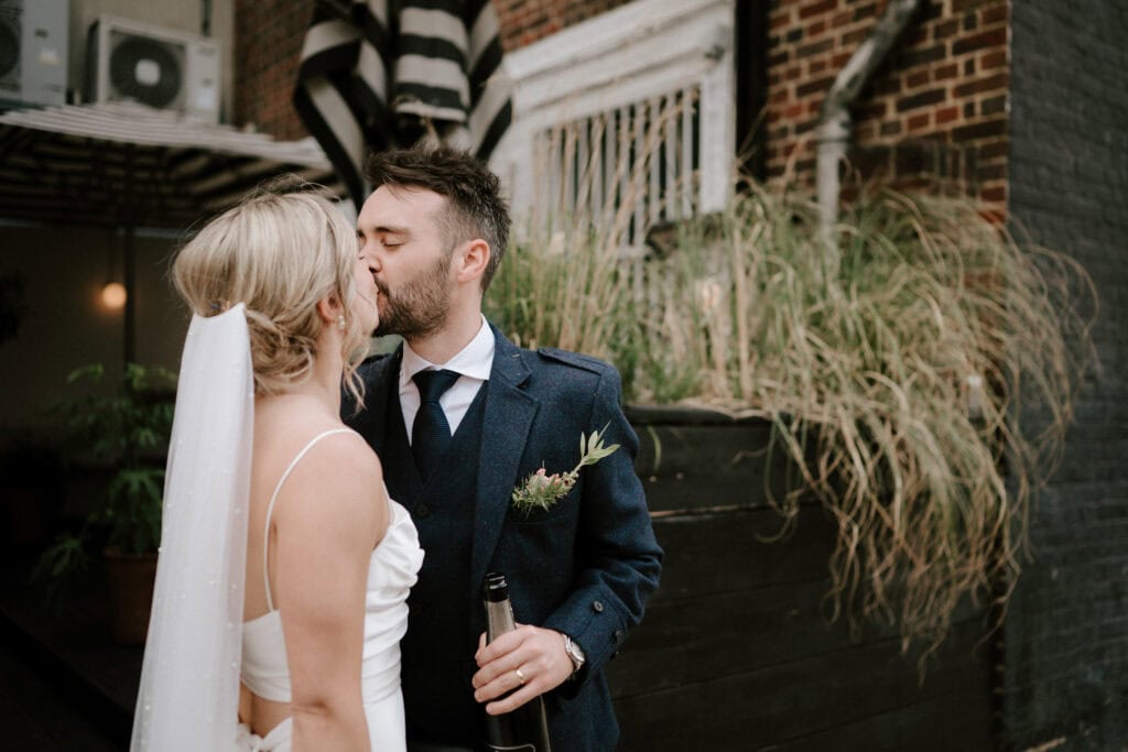 A bride and groom share a kiss outdoors, captured beautifully by a Bethnal Green wedding photographer. The bride is in a white dress and veil, while the groom sports a dark suit and tie, holding a bottle. They stand in front of a brick building with plants and a striped awning in the background. Image by Pearce Wedding Photography.