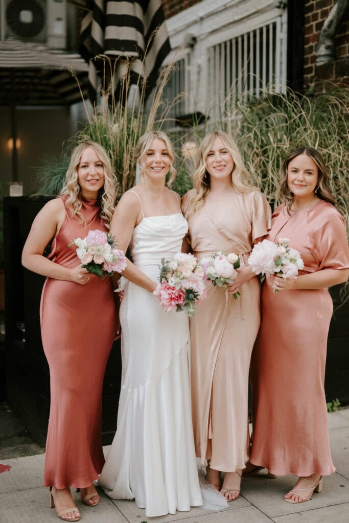 Four women smile and stand side by side. The bride, in a white dress, is third from the left. The three bridesmaids wear different shades of pink and gold dresses, each holding a bouquet of flowers. They are outdoors, with tall grass and a building in the background—captured perfectly by a Bethnal Green wedding photographer. Image by Pearce Wedding Photography.