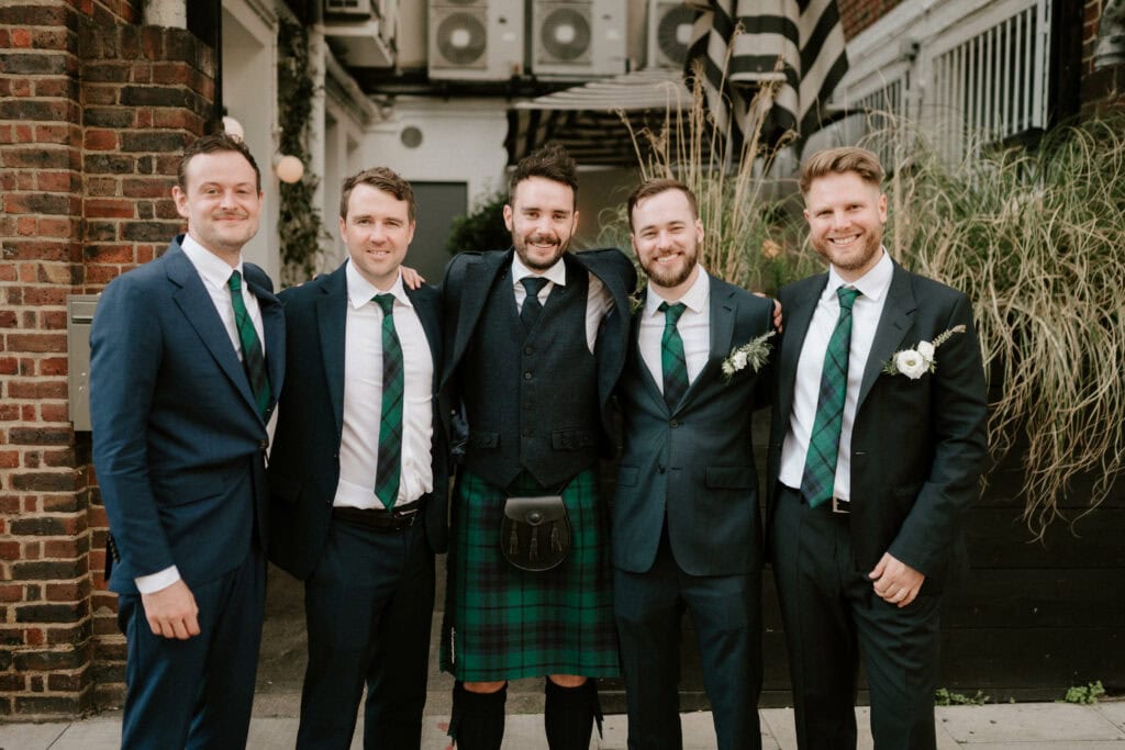 Five men in formal attire pose together outside. Four wear dark suits with white shirts and green ties, while the man in the center wears a green tartan kilt with a dark jacket. Captured by a Bethnal Green wedding photographer, they stand in front of a brick building with green plants and air conditioning units visible behind them. Image by Pearce Wedding Photography.