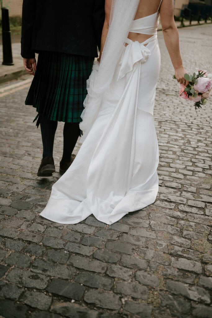 A bride and groom walk down a cobblestone street. The bride, in a flowing white gown with a bow at the back, holds a bouquet of roses. The groom, dressed in a green plaid kilt, walks beside her. Captured by a Bethnal Green wedding photographer, the focus is on their attire and the charming street. Image by Pearce Wedding Photography.