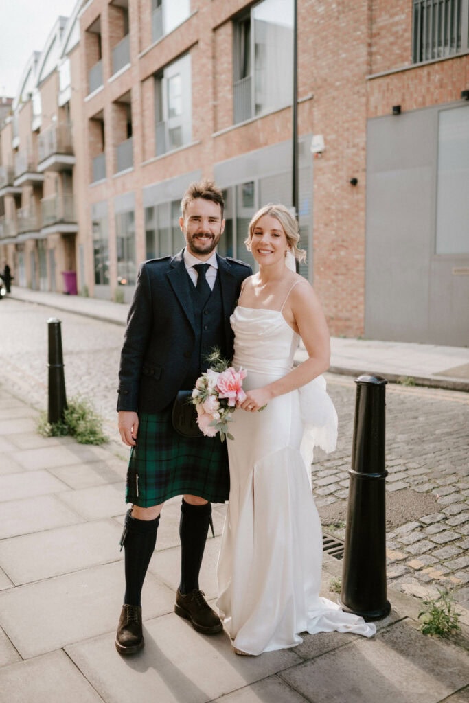 A couple posing for a photo on a city street. The man is wearing traditional Scottish attire, including a kilt, and the woman is dressed in a white wedding gown, holding a bouquet of flowers. They are both smiling, standing in front of a modern brick building, captured beautifully by their Bethnal Green wedding photographer. Image by Pearce Wedding Photography.