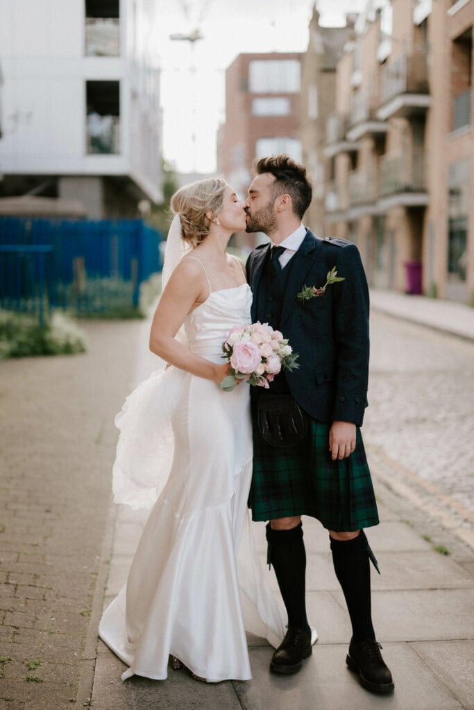 A bride and groom share a kiss on a cobblestone street. The bride is in a white strapless wedding gown with a long veil, holding a bouquet of flowers. The groom is dressed in a traditional Scottish kilt outfit. Urban buildings serve as their backdrop, beautifully captured by their Bethnal Green wedding photographer. Image by Pearce Wedding Photography.