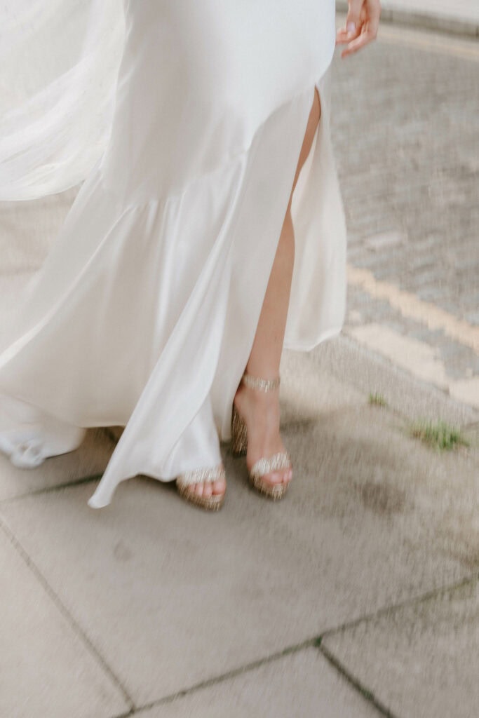 A bride in a white dress walking on a sidewalk, showing a partial view of her gown with a slit revealing her leg and feet in sparkling high-heeled shoes, perfectly captured by a Bethnal Green wedding photographer. The pavement and a small patch of grass are visible in the background. Image by Pearce Wedding Photography.