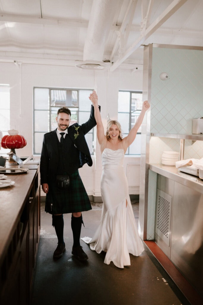 A couple celebrate joyfully indoors. The woman is wearing a white wedding dress with raised arms, and the man is sporting a dark suit with a kilt, also raising one arm. They are smiling brightly, captured perfectly by their Bethnal Green wedding photographer, with kitchen-like surroundings in the background. Image by Pearce Wedding Photography.