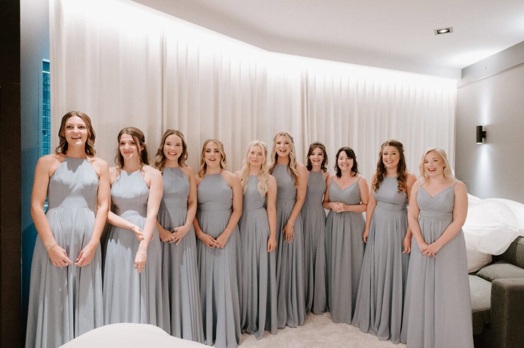 A group of nine women in light blue dresses stand together in a well-lit room at The Cave Hotel in Kent, smiling at the camera, ready for the wedding celebration.