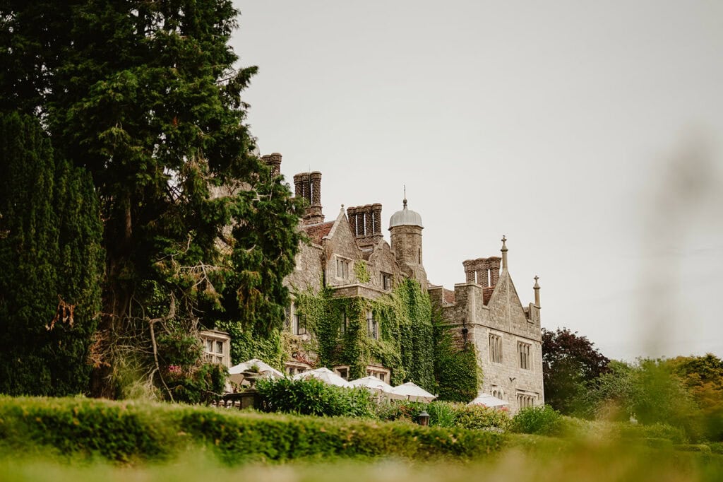 A historic, ivy-covered stone building with multiple chimneys and dormer windows, surrounded by lush greenery and trees on an overcast day, reminiscent of Eastwell Manor in Kent—a perfect setting for a charming wedding.
