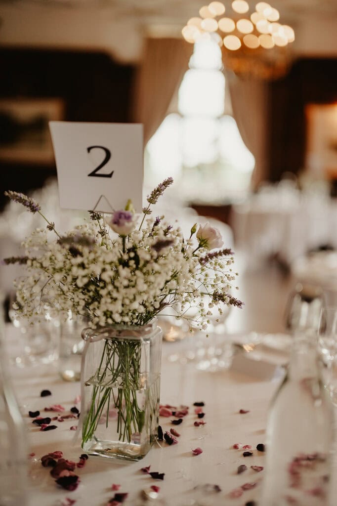 A wedding reception table at Eastwell Manor featuring a glass vase with white and purple flowers, a table number card labeled "2," and scattered flower petals. In the background, more tables and a chandelier add to the enchanting Kent setting.