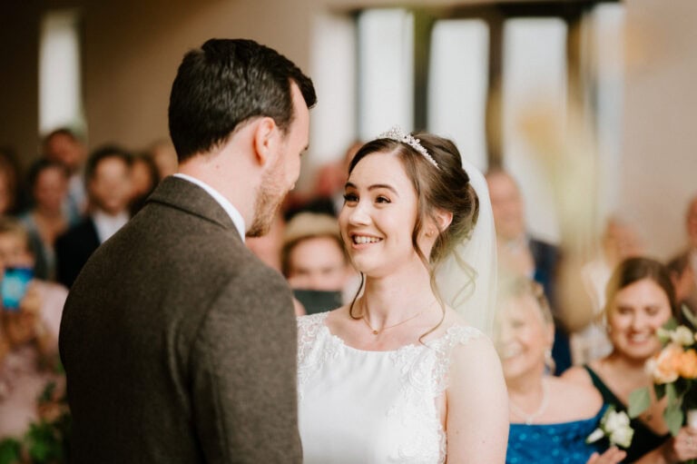A bride and groom smile at each other during their wedding ceremony, with the bride wearing a white dress and tiara and the groom in a brown suit. Guests seated in the background look on, smiling and holding flowers. Image by Pearce Wedding Photography.