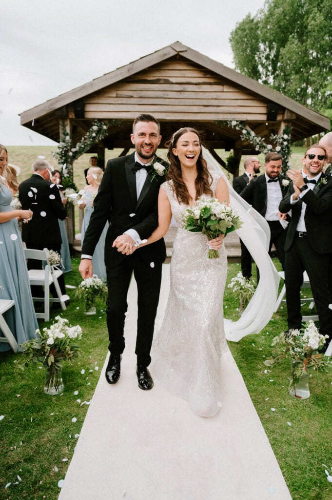 A married couple, holding hands, walks down an aisle outdoors at the Cave Hotel in Kent. The bride is in a white dress holding a bouquet, and the groom is in a black suit and bow tie. Guests are clapping and throwing confetti.