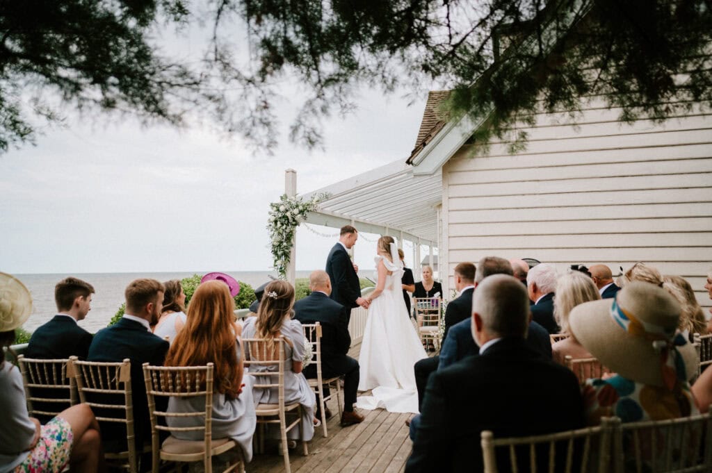 A couple stands at the altar during an outdoor wedding ceremony by the water at Beacon House in Kent, surrounded by seated guests on wooden chairs.