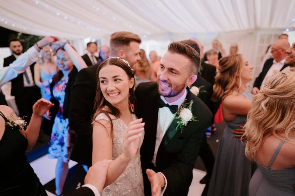 A bride and groom wearing formal attire smile and dance together at a wedding reception, surrounded by guests under a canopy with string lights at The Cave Hotel in Kent.