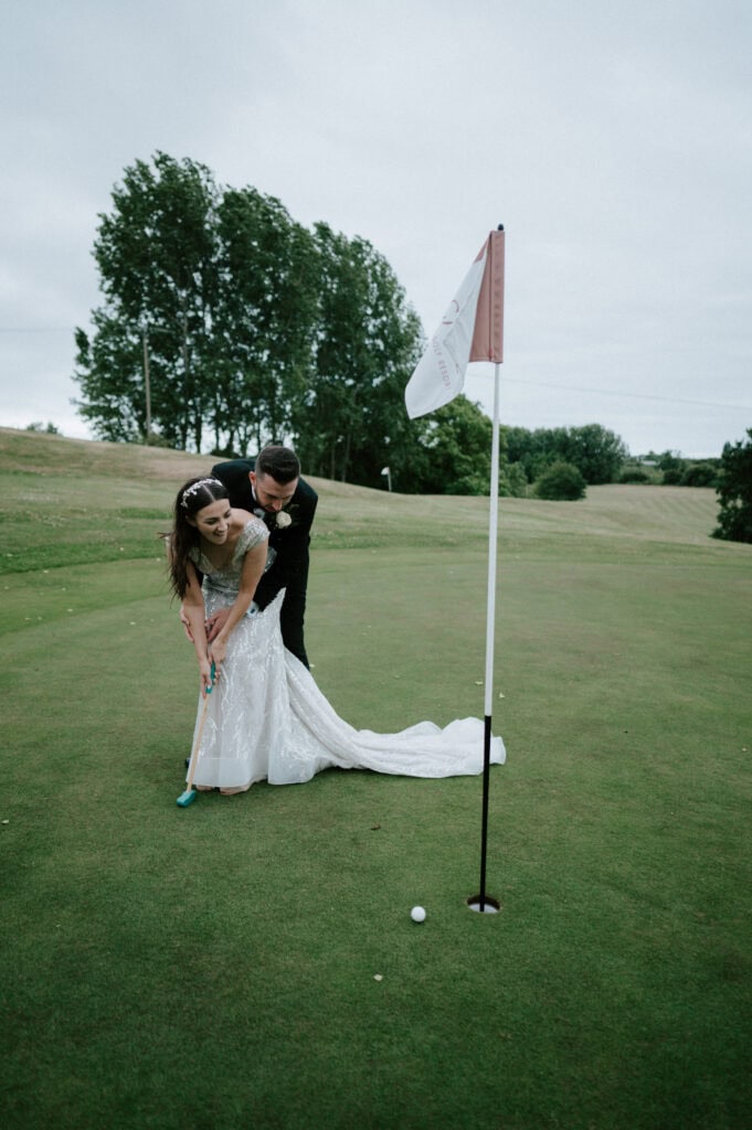 At The Cave Hotel in Kent, a bride in a wedding dress and a groom in a suit playfully putt a golf ball on the green, with a flagstick and trees providing the perfect wedding backdrop.