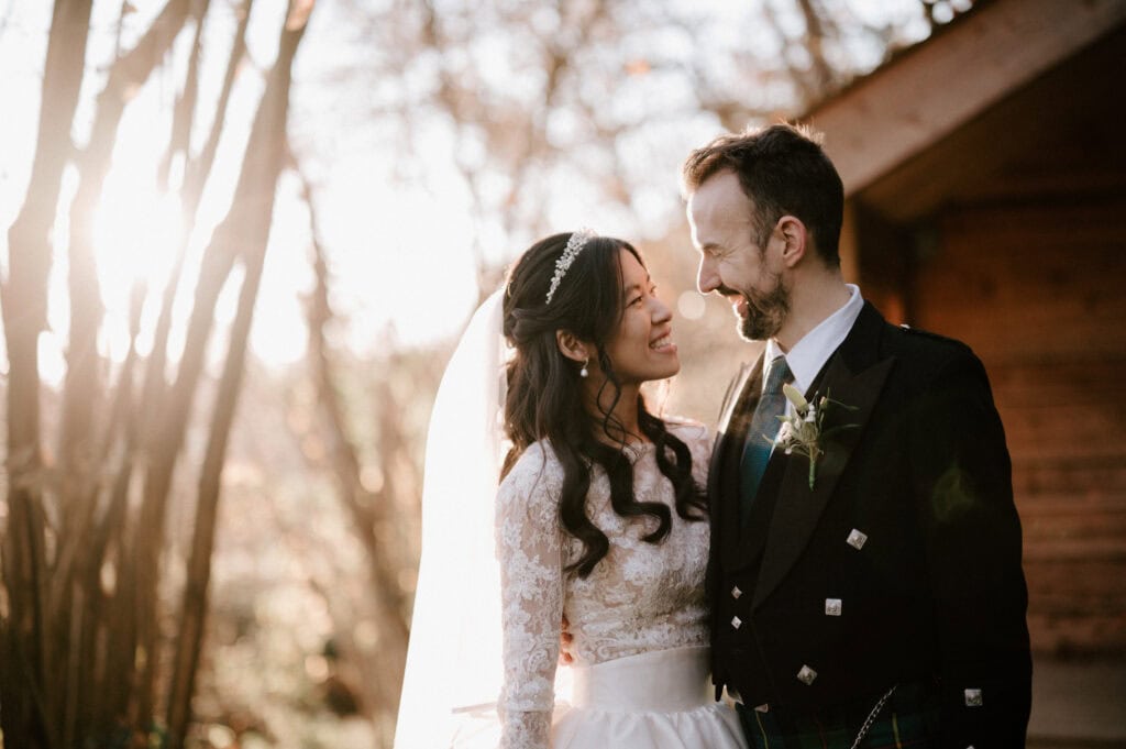 At their enchanting Millbridge Court wedding, the bride beams in a lace dress and veil at the groom, who sports a traditional Scottish kilt and suit. They stand outdoors, with the sun casting a gentle glow through the trees behind them. Image by Pearce Wedding Photography.
