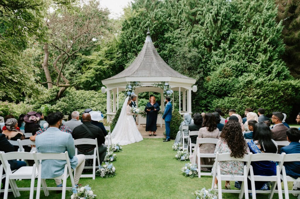 A wedding ceremony is taking place outdoors in beautiful Maidstone. The bride and groom stand under a white gazebo adorned with flowers, facing the officiant. Guests are seated in white chairs on either side of the aisle, surrounded by lush greenery and trees. Image by Pearce Wedding Photography.
