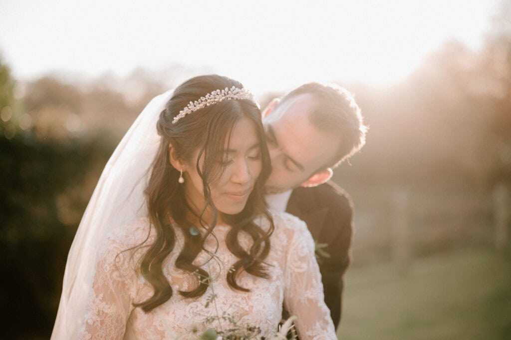 At Millbridge Court, a bride and groom share an intimate moment outdoors at sunset. The bride, in a lace wedding dress and pearl headband, smiles softly as the groom gently kisses her forehead. They are surrounded by a peaceful, natural setting that beautifully enhances their wedding day. Image by Pearce Wedding Photography.