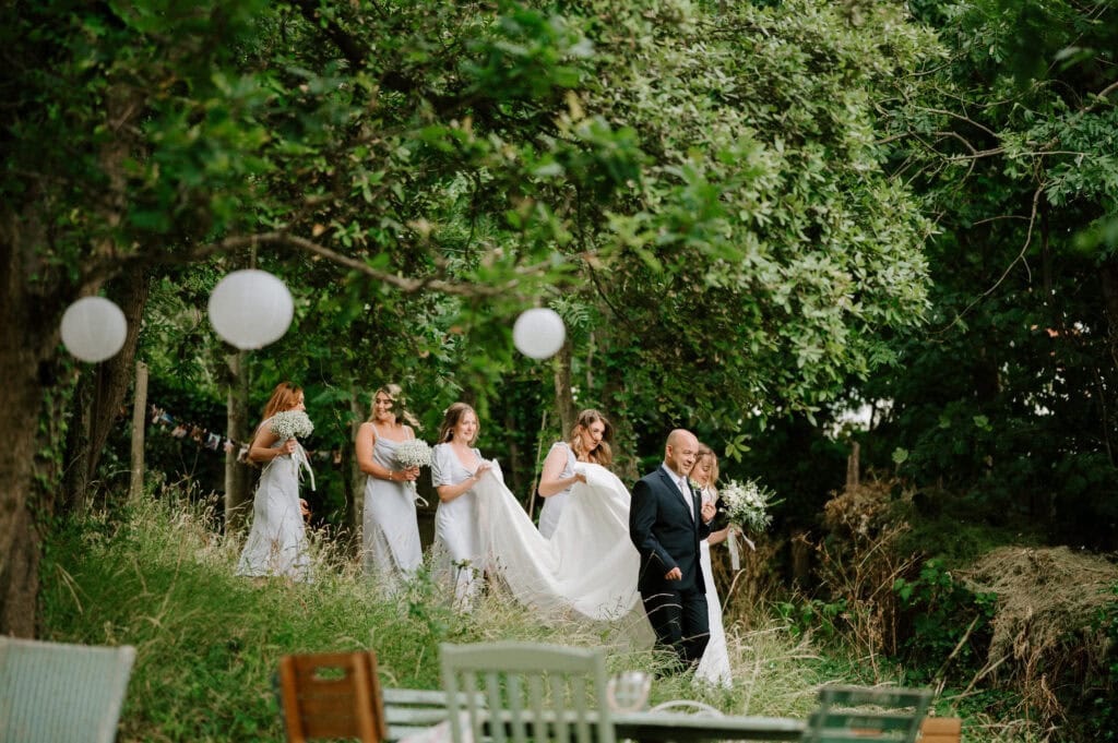 A bride and five bridesmaids, all wearing white dresses, walk downhill on a grassy path among trees at the picturesque Beacon House in Kent. A man in a dark suit leads them as white spherical decorations hang from the trees, adding to the enchanting wedding ambiance.
