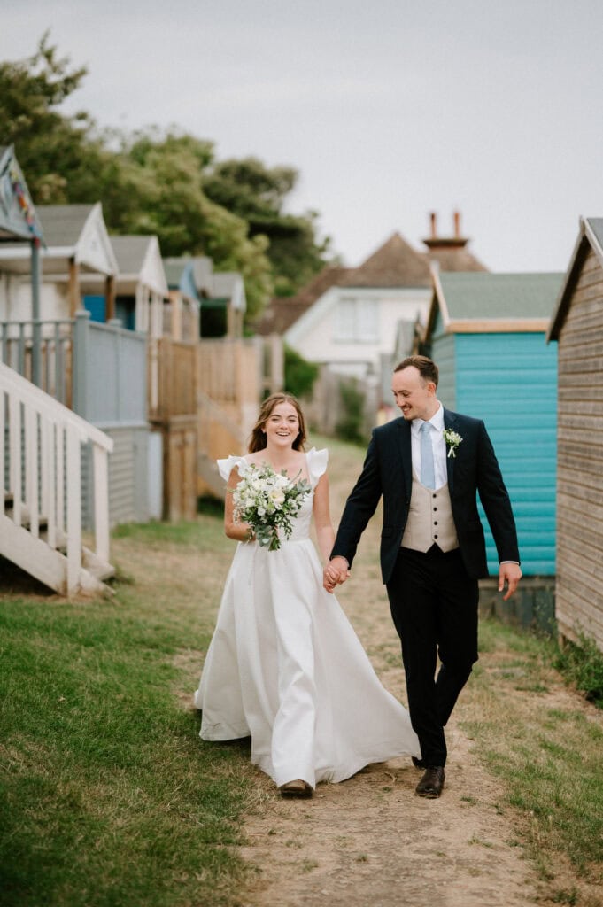 A bride and groom walk hand in hand along a grassy path between beach huts near Beacon House in Kent. Both are smiling, the bride holding a bouquet of white flowers.
