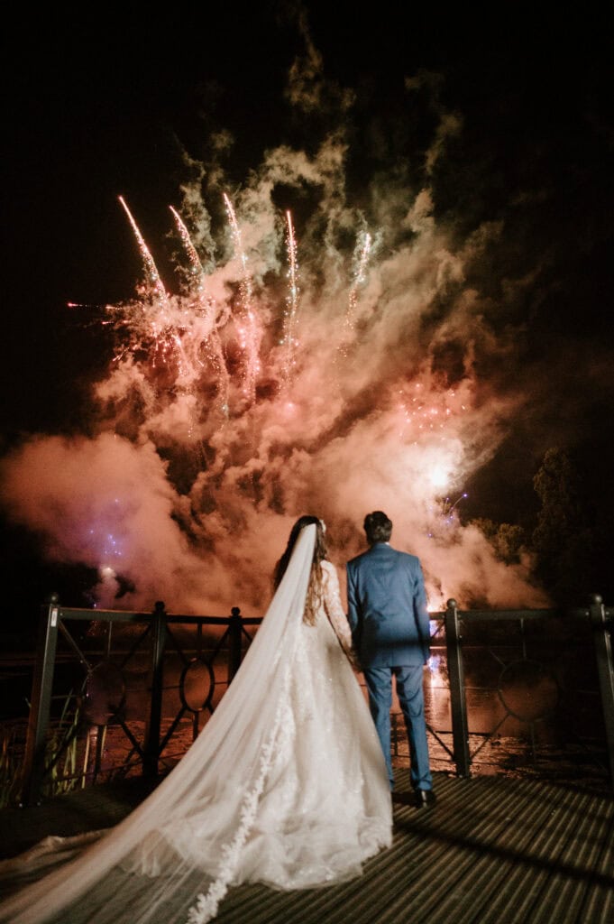 A newlywed couple stands on a bridge at night, viewing colorful fireworks illuminating the sky. Captured by a talented Kent wedding photographer, the bride is in a long white dress with a flowing veil, and the groom in a blue suit. Smoke adds dramatic effect to this vibrant scene. Image by Pearce Wedding Photography.