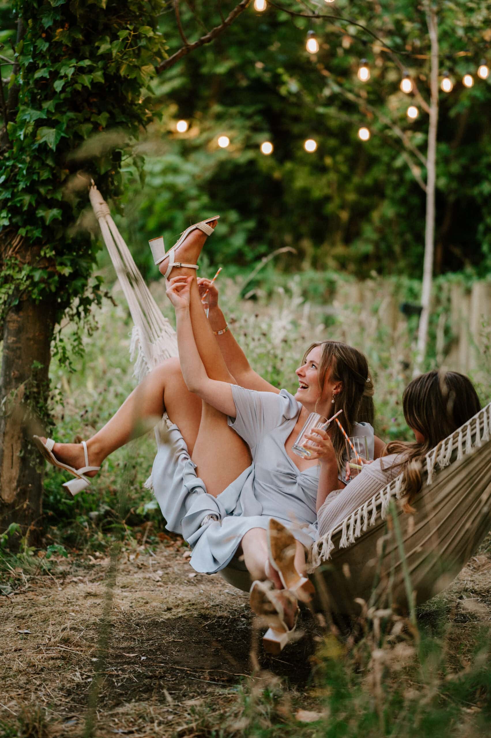 Two women laughing and relaxing on a hammock in a lush garden, captured by a talented Kent wedding photographer. They hold drinks, playfully kicking their legs as string lights above create a cozy, festive atmosphere. Image by Pearce Wedding Photography.