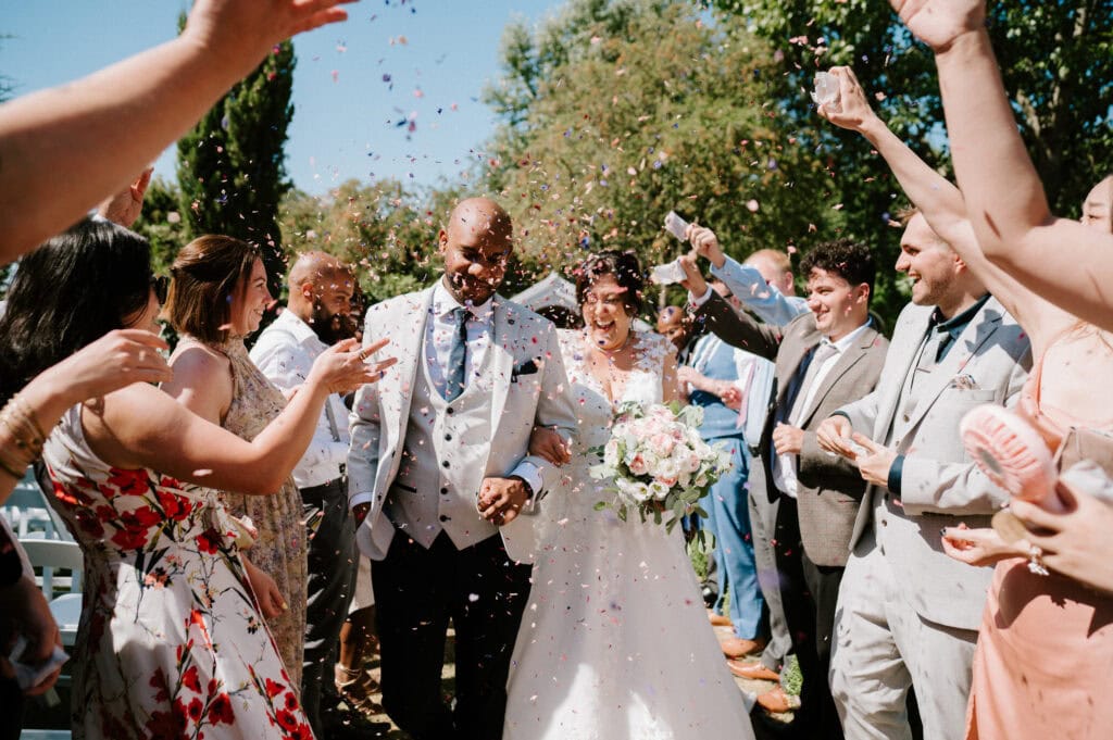 A bride and groom walk down the aisle outdoors at Marleybrook House in Kent while guests throw confetti and celebrate. The bride is holding a bouquet, and both are smiling.