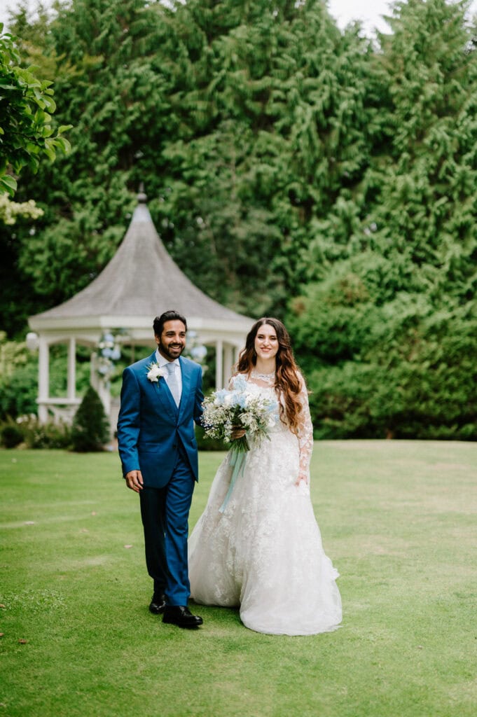A bride and groom stand on a well-manicured lawn in front of a charming gazebo at The Orangery in Kent. The bride, in her stunning white dress, holds a bouquet, while the groom looks dapper in a blue suit. Lush trees provide a beautiful backdrop to this picturesque wedding scene.