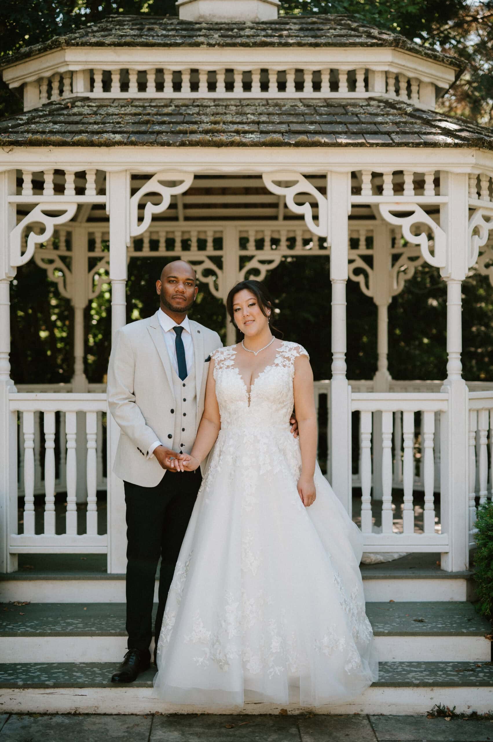 A couple stands in front of a white gazebo, holding hands, on their wedding day. The person on the left is wearing a light grey suit with a dark tie, while the person on the right is in a white lace wedding gown. Both are smiling subtly, captured beautifully by their Kent wedding photographer amidst lush greenery. Image by Pearce Wedding Photography.