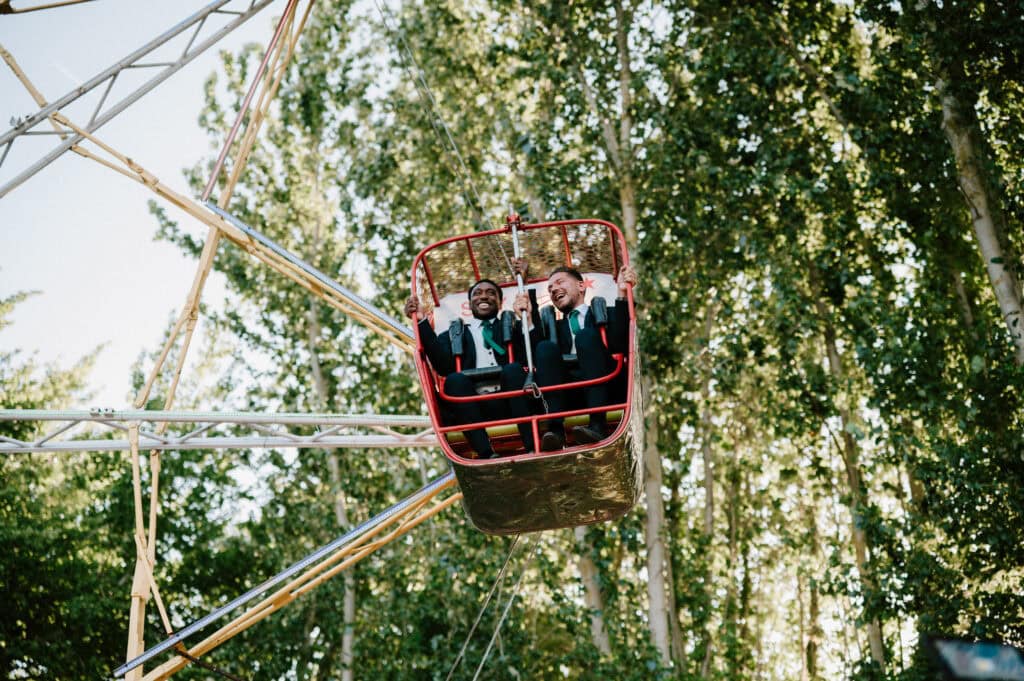 Two people dressed in formal attire are smiling as they ride in a Ferris wheel at Marleybrook House, with tall, leafy trees providing a picturesque backdrop on their wedding day.