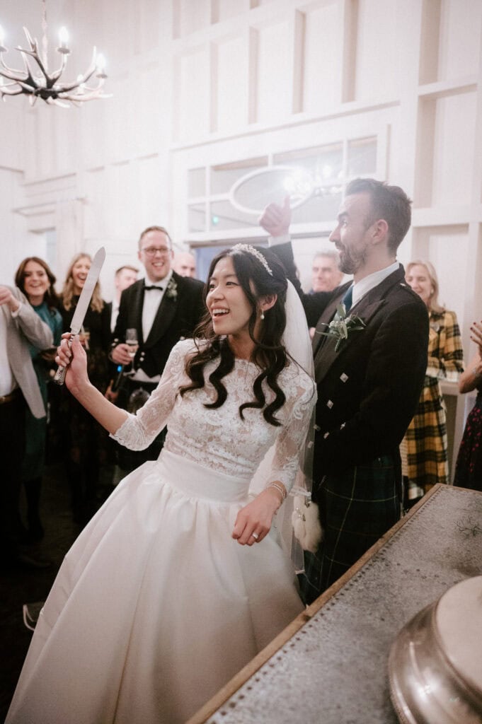 A bride, wearing a white wedding dress and veil, joyfully holds a cake knife while standing next to the groom in a kilt. They are surrounded by smiling and applauding guests in formal attire, set in an elegant venue with chandeliers. Image by Pearce Wedding Photography.