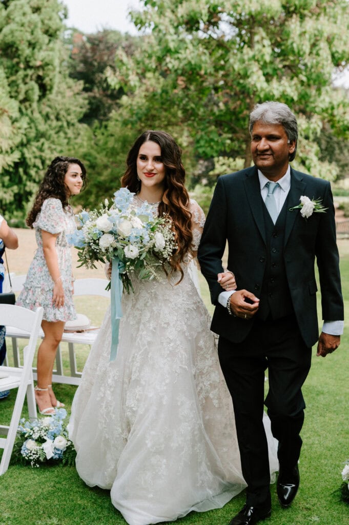 A bride in a white lace gown holds a blue and white bouquet, walking arm in arm with an older man in a suit down an outdoor aisle at The Orangery in Kent, surrounded by seated guests and trees.