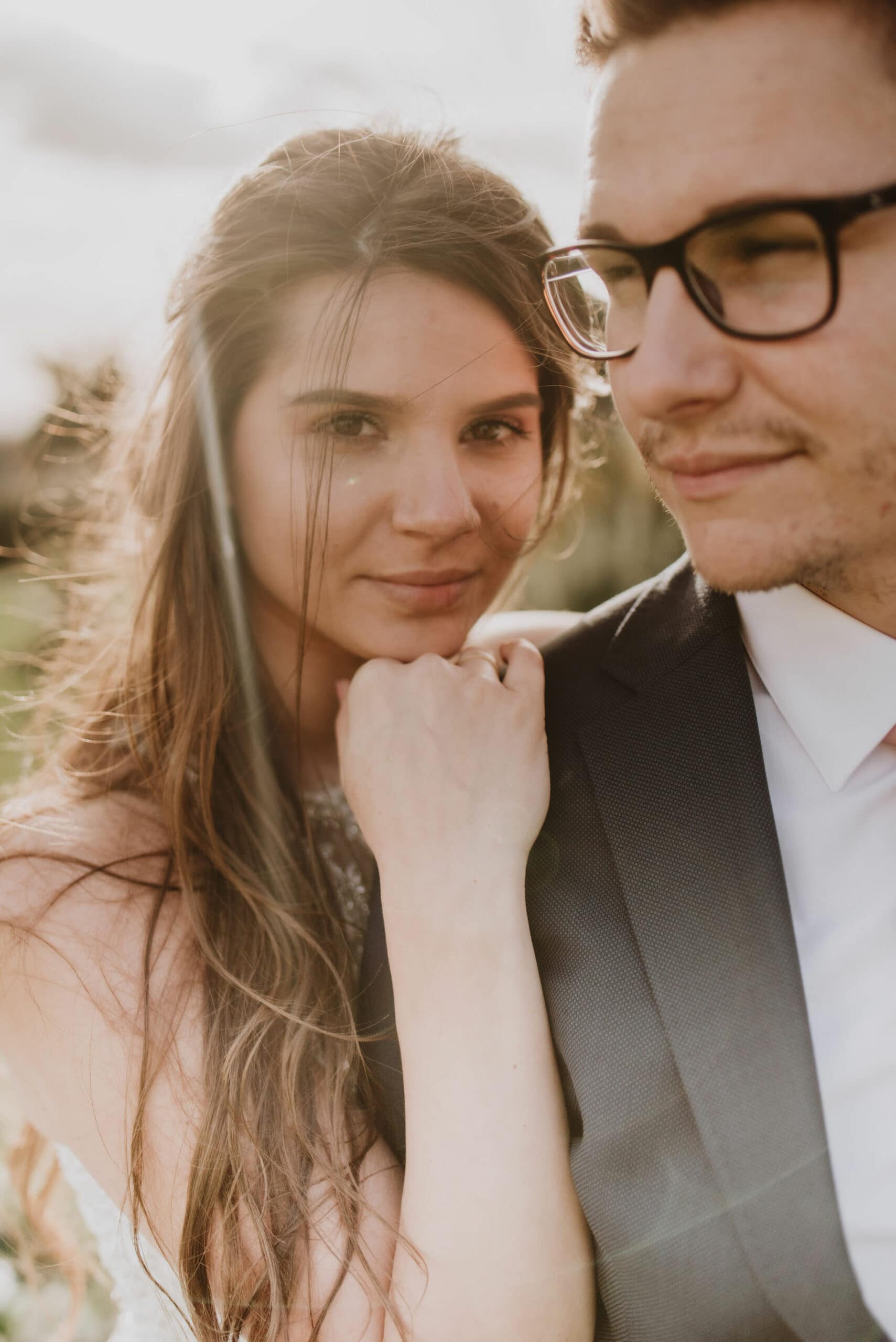 A woman with long brown hair and a man wearing glasses and a suit stand close together in an outdoor setting at Albion House. The woman has a gentle smile and rests her chin on her hand. Sunlight softly illuminates them, creating a warm, intimate atmosphere, perfect for a Thanet wedding photographer to capture. Image by Pearce Wedding Photography.
