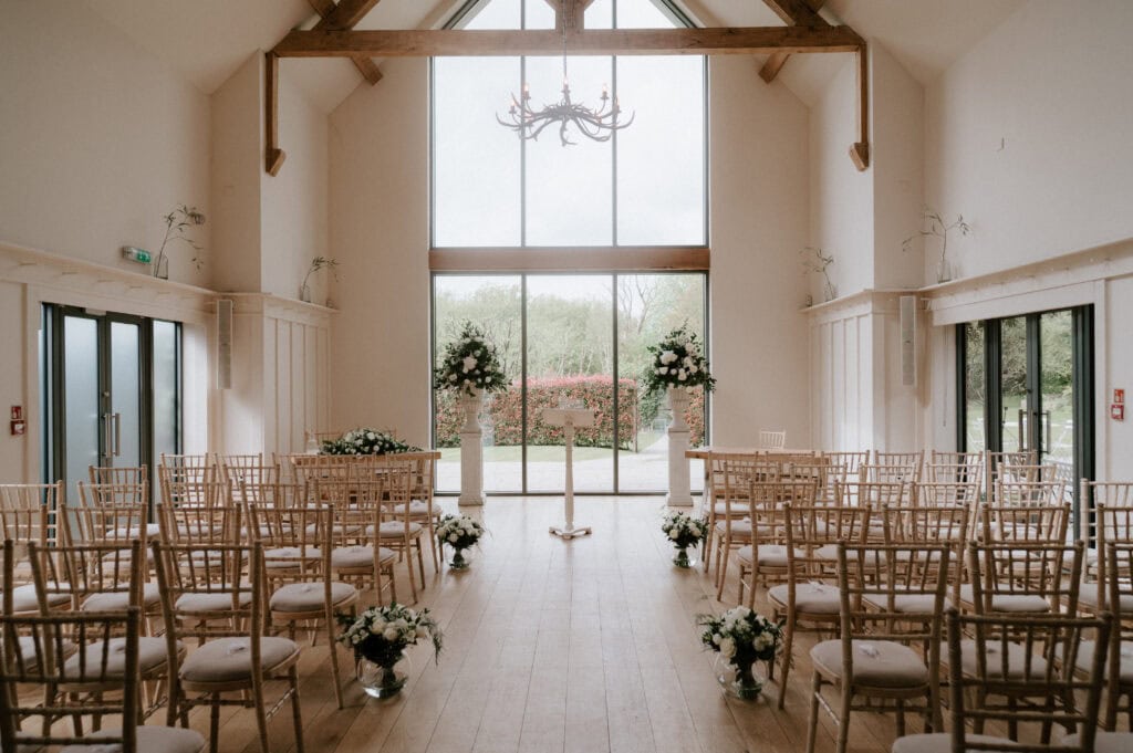 The beautifully decorated wedding ceremony space at Millbridge Court features a barn-style venue. Wooden chairs face a glass wall overlooking a lush green landscape, while floral arrangements with white flowers and greenery line the aisle. A chandelier graces the wooden ceiling beams. Image by Pearce Wedding Photography.