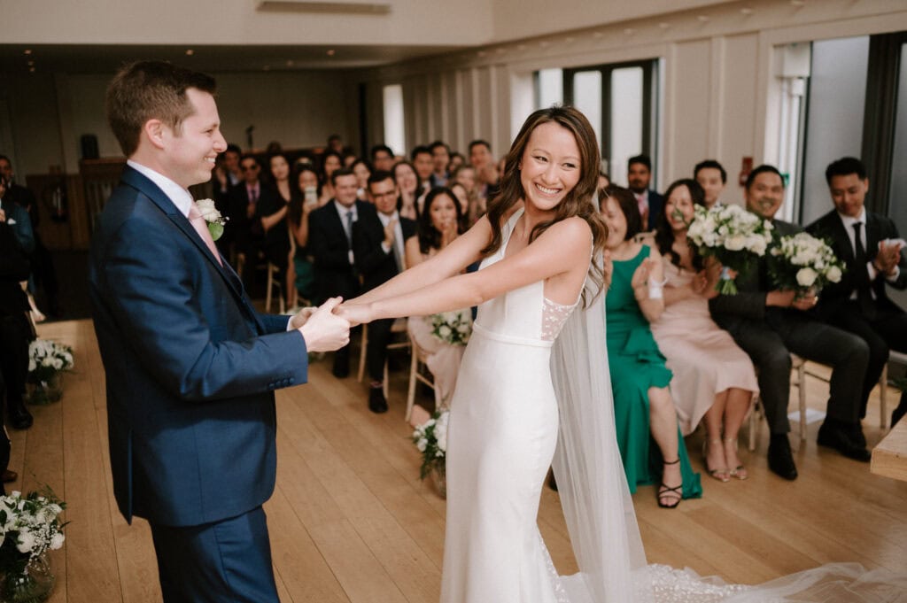 At Millbridge Court, a bride and groom hold hands and smile during their wedding ceremony. The bride wears a white gown with a long veil, while the groom looks dapper in a navy suit. Guests seated in the beautifully decorated room with white flowers clap joyfully. Image by Pearce Wedding Photography.