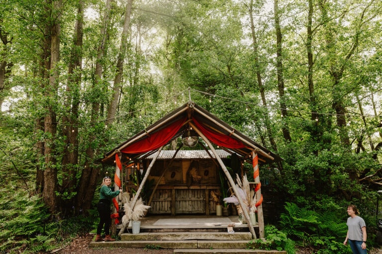 A tent-like wooden structure with red drapes is set in a lush, green forest, making it a unique wedding venue in Kent. Two people are nearby: one stands on the left holding a bucket, and the other walks on the right. Ferns and trees surround this picturesque spot, perfect for wilderness weddings. Image by Pearce Wedding Photography.