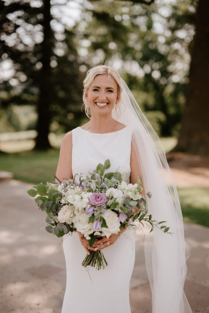 A smiling bride in a white wedding dress and veil stands outdoors at Highley Manor, holding a bouquet of white and lavender flowers with greenery. She has a light complexion and blonde hair, with trees and greenery blurred in the background. Image by Pearce Wedding Photography.