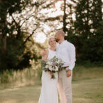 A couple stands closely together outdoors with trees in the background, smiling warmly. The woman, in a white dress and holding a bouquet of flowers, leans into the man dressed in a white shirt and light-colored pants, who wraps an arm around her shoulder. This moment at Highley Manor feels magical. Image by Pearce Wedding Photography.
