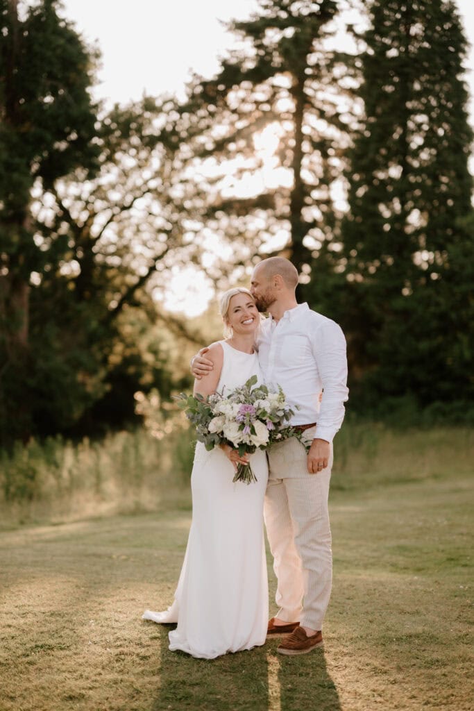 A couple stands closely together outdoors with trees in the background, smiling warmly. The woman, in a white dress and holding a bouquet of flowers, leans into the man dressed in a white shirt and light-colored pants, who wraps an arm around her shoulder. This moment at Highley Manor feels magical. Image by Pearce Wedding Photography.