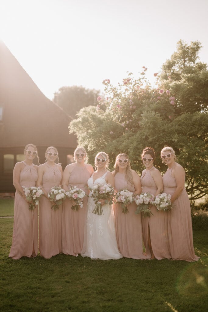 A bride in a white dress and six bridesmaids in matching blush pink dresses and sunglasses stand together outdoors at The Old Kent Barn, holding bouquets of pink and white flowers. They are all smiling, with greenery and flowers in the background, making for a perfect wedding scene. Image by Pearce Wedding Photography.