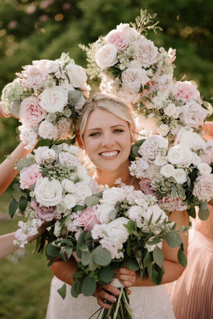 A smiling woman with blonde hair is surrounded by an arrangement of pink and white floral bouquets, held by people standing around her. She is outdoors at The Old Kent Barn, with a backdrop of greenery and soft sunlight filtering through the leaves, capturing the joy of a beautiful wedding day. Image by Pearce Wedding Photography.