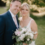 A smiling couple on their wedding day stand close together outdoors at The Old Kent Barn. The groom is wearing a dark blue suit with a light pink tie, and the bride is in a white lace dress holding a bouquet of flowers. The background is green and sunny, with blurred foliage. Image by Pearce Wedding Photography.