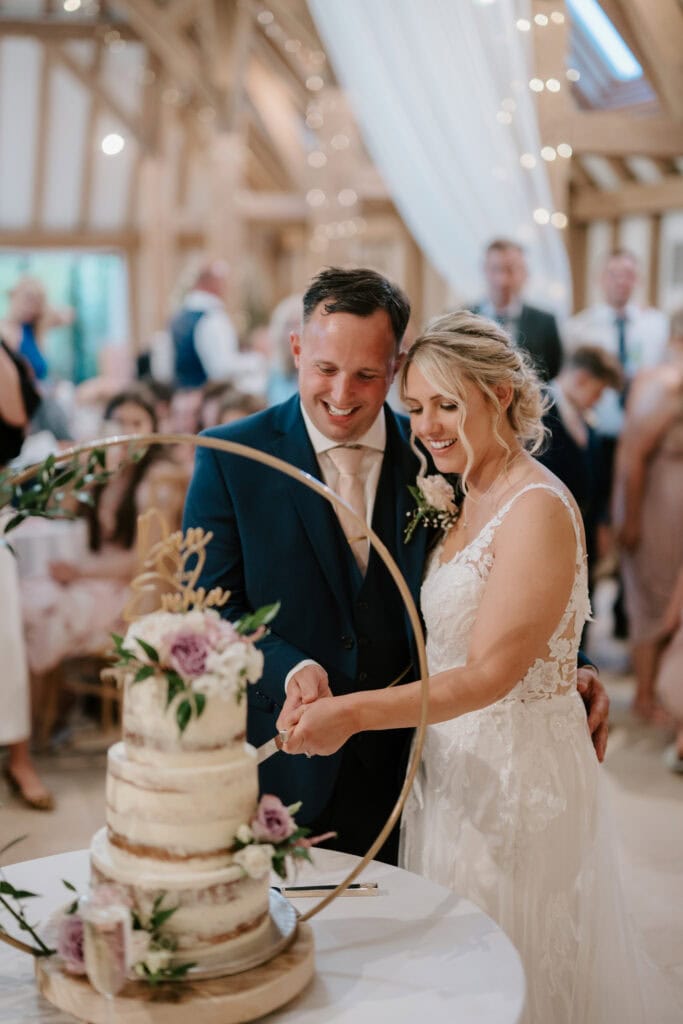 A bride and groom smile joyfully as they cut their wedding cake together. The cake, a three-tiered semi-naked design adorned with purple and white flowers, stands beautifully at The Old Kent Barn. Guests in the background look on, seated and standing in the warmly lit wedding reception venue. Image by Pearce Wedding Photography.