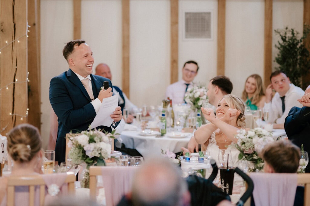 Groom giving speech at wedding, bride smiling and laughing.