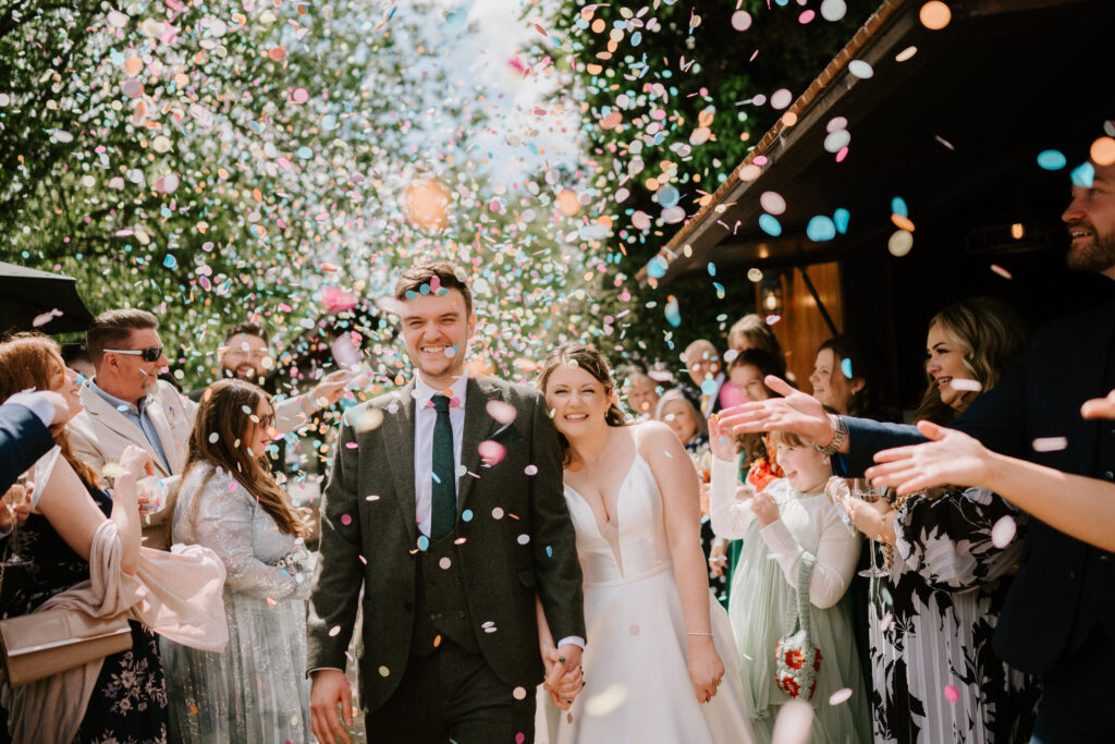 Newlyweds smiling under confetti surrounded by guests.