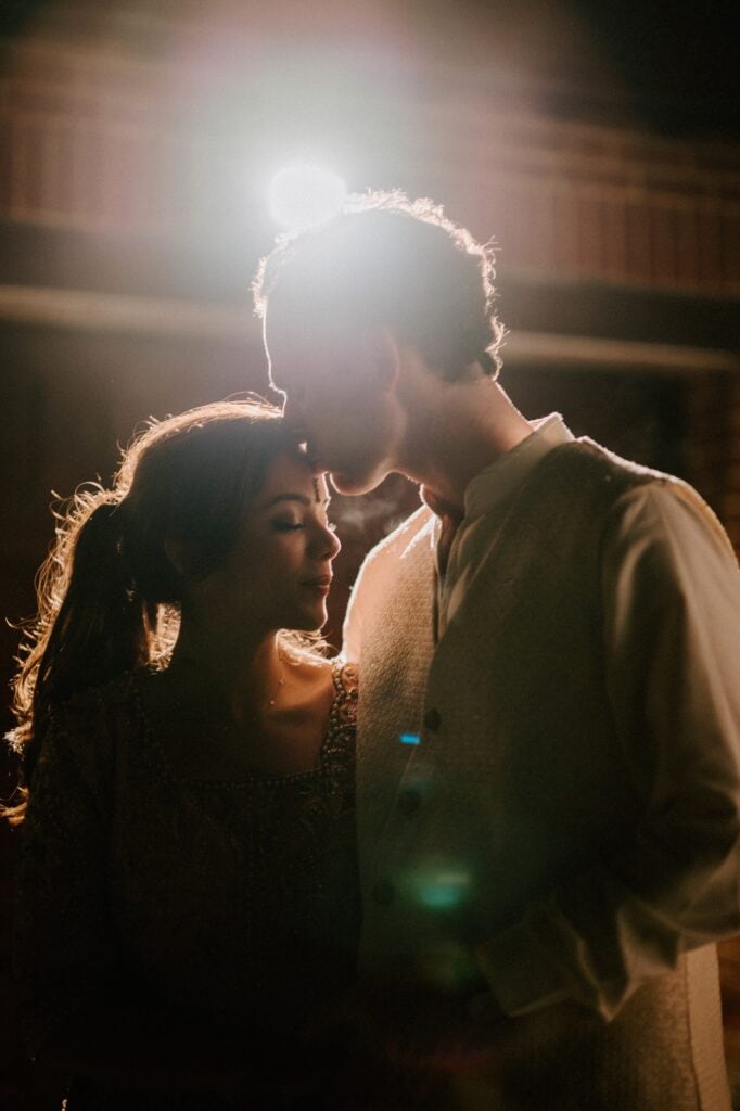 A couple stands closely in dim lighting, the man gently kissing the woman's forehead. Soft, warm light creates a romantic atmosphere captured by a wedding photographer in Kent, highlighting their peaceful expressions and casting a gentle glow around them. Image by Pearce Wedding Photography.