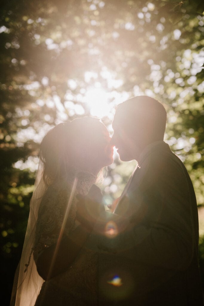 A bride and groom share a kiss in a sun-dappled forest setting, beautifully captured by a Kent wedding photographer. The sunlight creates a warm glow around them, highlighting the bride's veil and the groom's silhouette. Image by Pearce Wedding Photography.