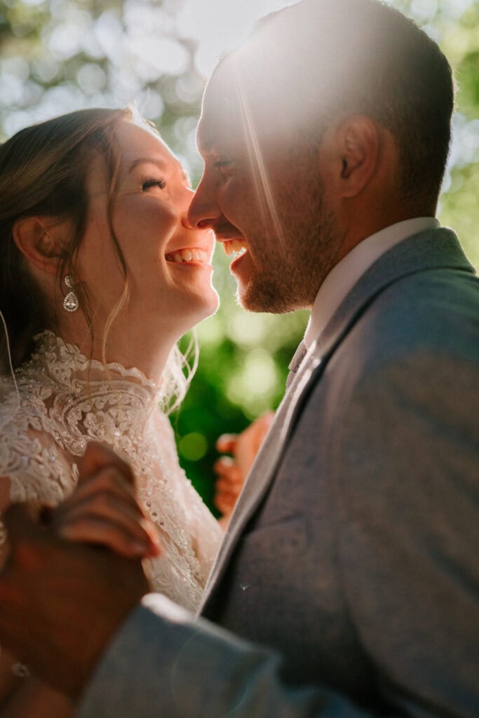 The bride and groom exchange smiles while holding hands outdoors at The Plough at Leigh. She wears a lace wedding dress and earrings, and he's in a light gray suit. Sunlight softly illuminates their joyous wedding moment. Image by Pearce Wedding Photography.